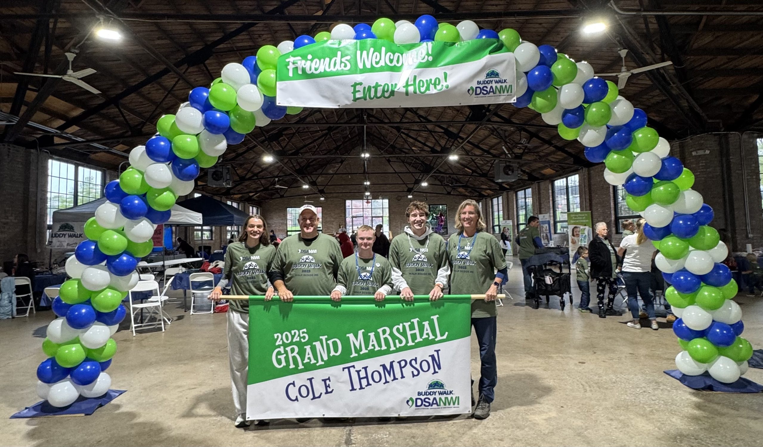 Cole Thompson and his team holding the 2025 Grand Marshal banner under the balloon arch at the Buddy Walk